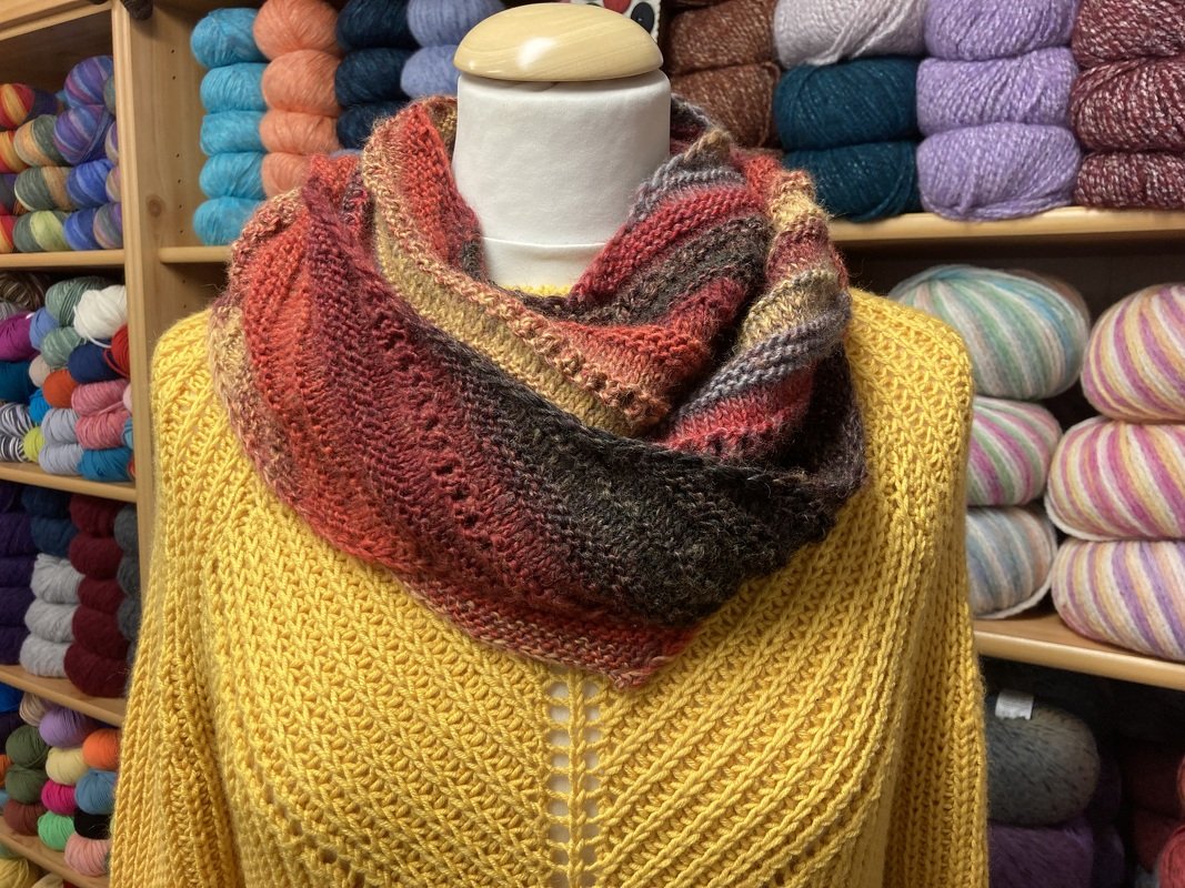 Mehrfarbiges gestricktes Dreieckstuch in Rot-, Braun- und Gelbtönen auf Schaufensterpuppe mit gelbem Pullover, Garnregale im Hintergrund