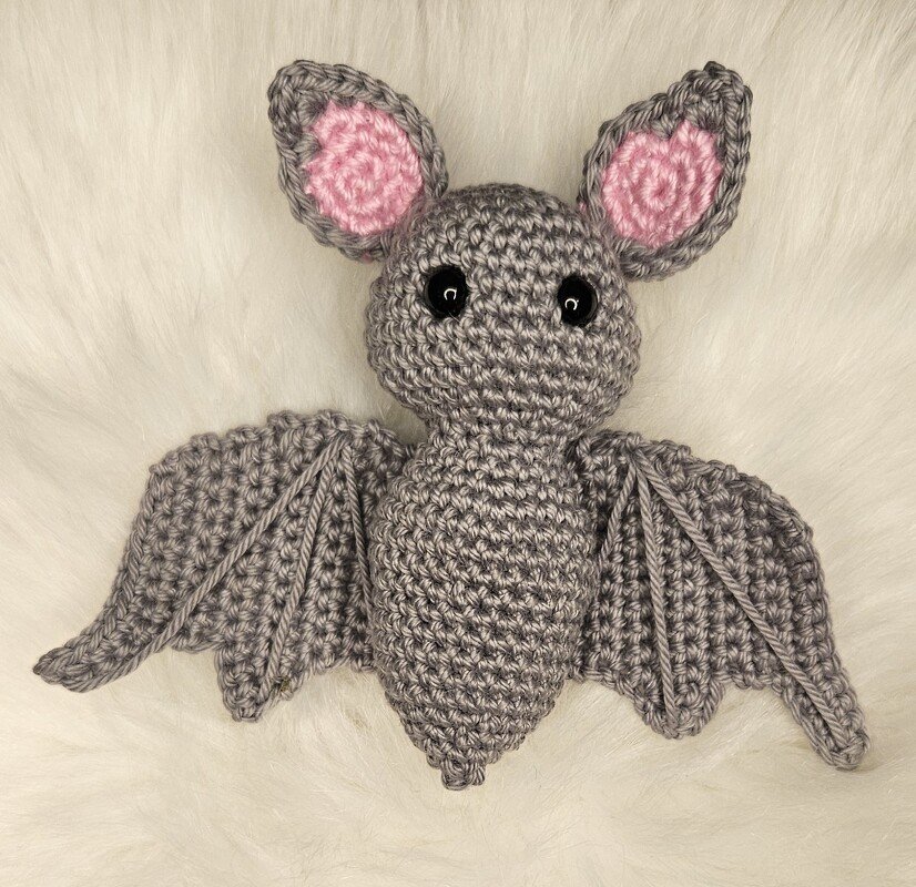 Gehäkelte graue Fledermaus‑Amigurumi mit rosa Ohren und schwarzen Augen auf weißem Kunstfell