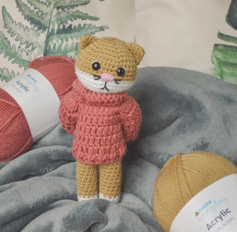Gehäkelte Amigurumi-Katze mit rosa Pullover zwischen Wollknäueln auf grauer Decke