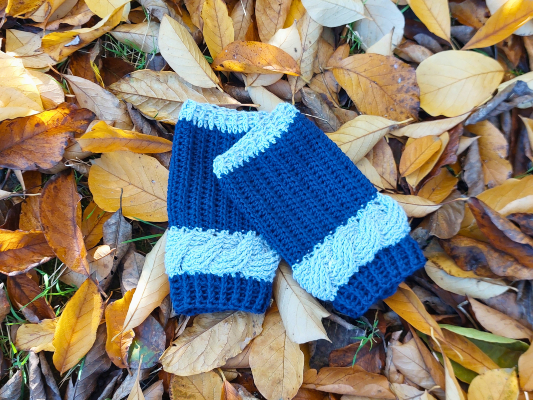 Zwei blaue gehäkelte Armstulpen mit Zopfmuster auf gelb-braunem Herbstlaub.