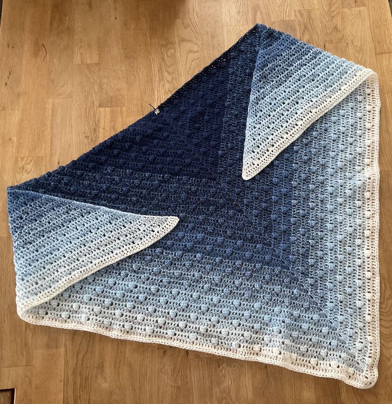 Ausgebreitetes gehäkeltes Dreieckstuch in Blauverläufen und Weiß mit Noppenstruktur, flach auf Holz liegend.