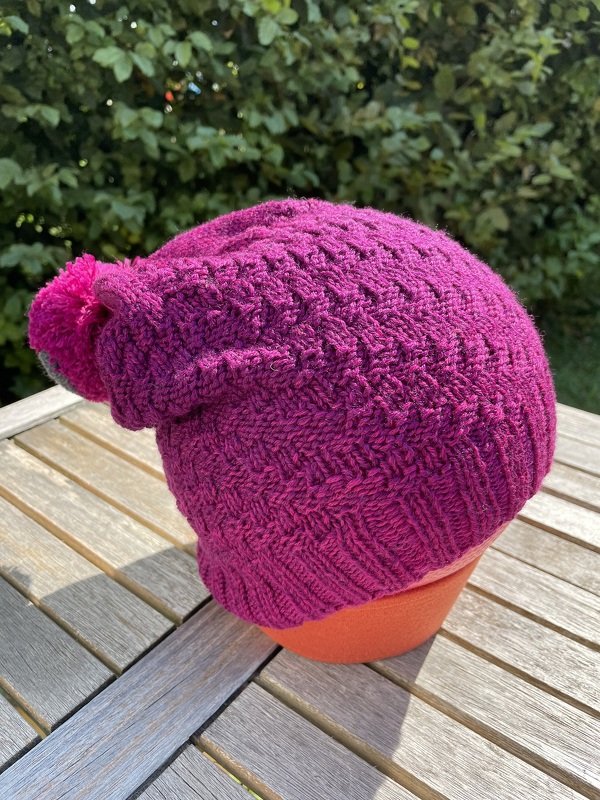 Fuchsiafarbene gestrickte Mütze mit Pompon auf orangefarbenem Kopfständer auf Holztisch, Heckenhintergrund