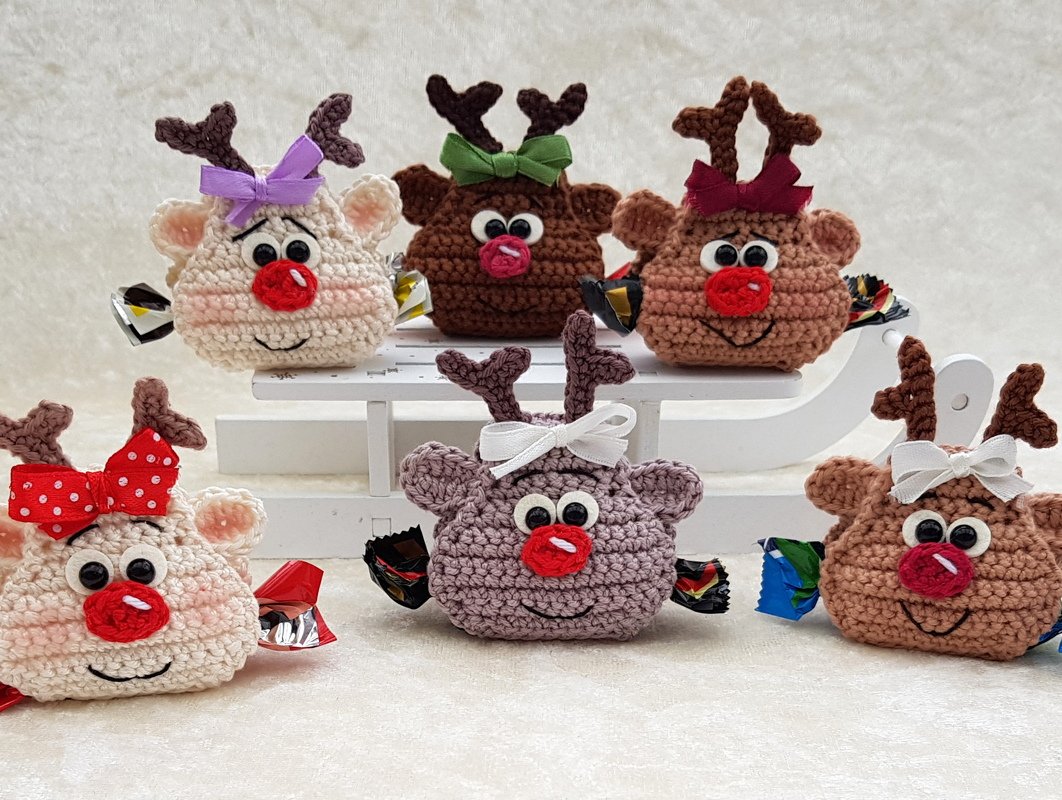 Sechs gehäkelte Rentiere mit roten Nasen und Schleifen, seitlich mit eingewickelten Süßigkeiten bestückt; teils auf weißem Schlitten.