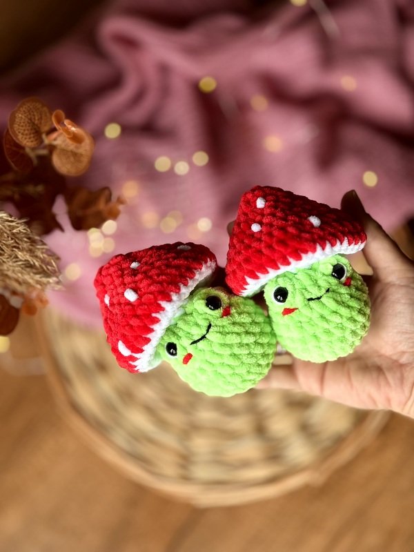 Two small green crochet mushroom plushies with red and white spotted caps held in a hand.