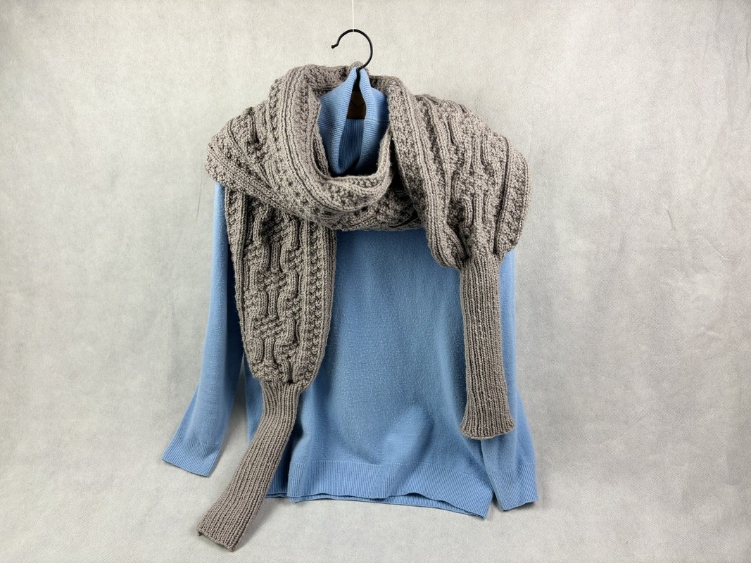 Taupefarbener, dicker Zopfmuster-Schal mit gerippten Enden über hellblauem Pullover an Kleiderbügel