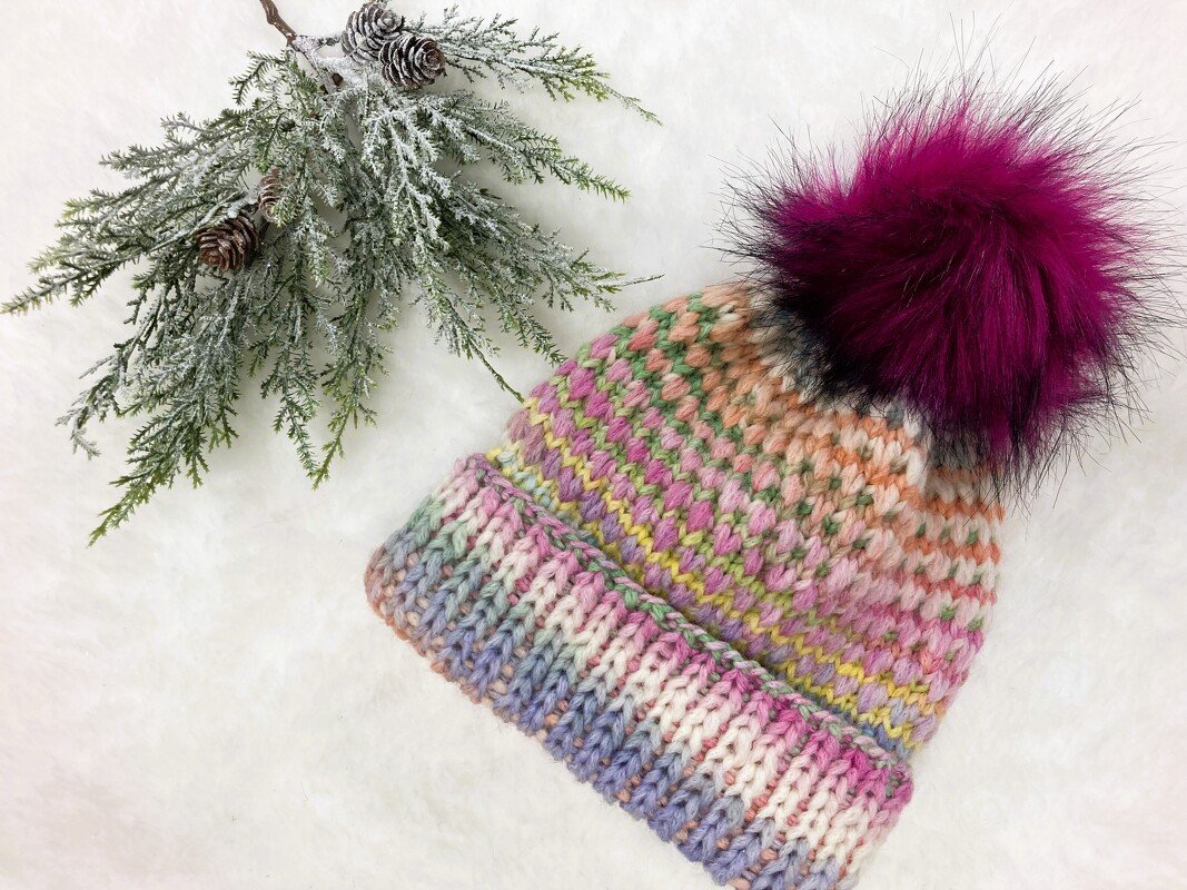 Bunte Strickmütze mit Rippenbündchen und dunkelpinkem Bommel auf weißem, flauschigem Untergrund, daneben ein gefrosteter Tannenzweig