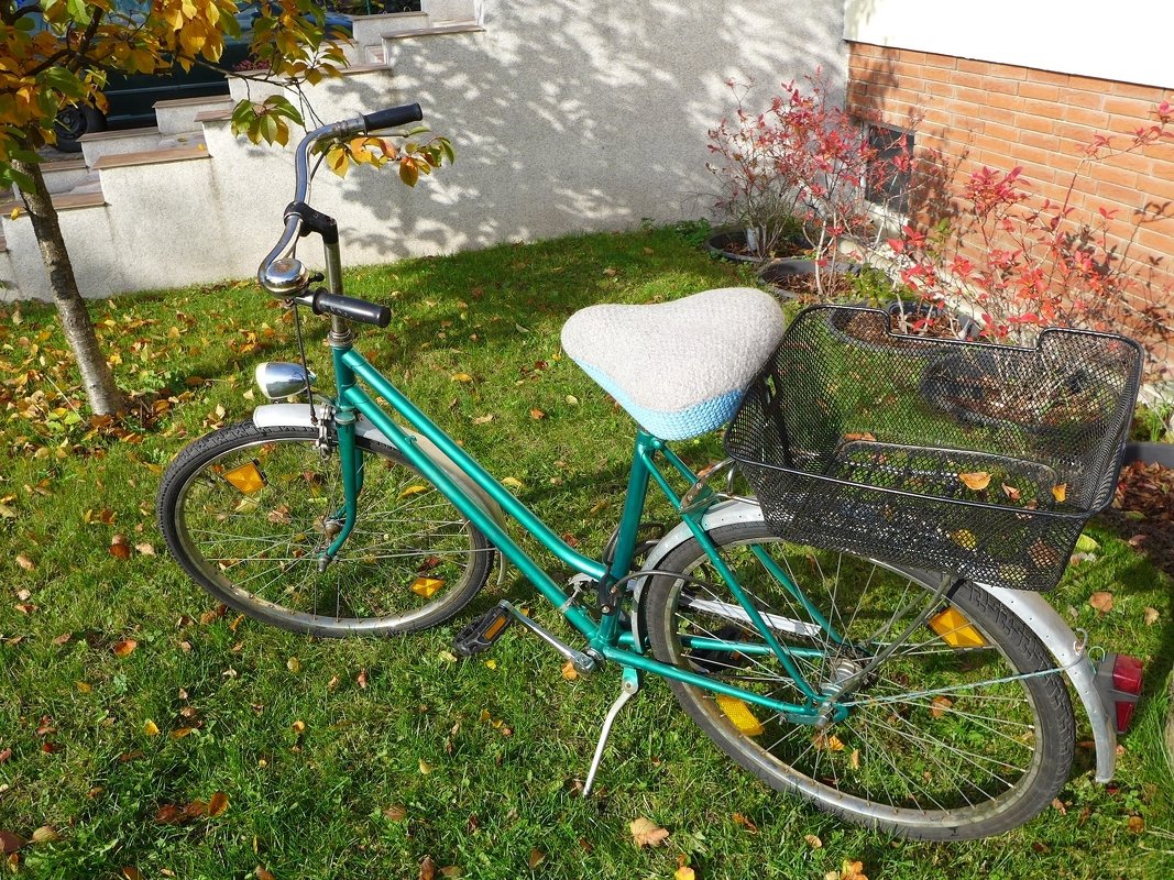 Türkisfarbenes Citybike mit hellgrauem, gestricktem Sattelbezug und hellblauem Rand sowie schwarzem Gepäckkorb