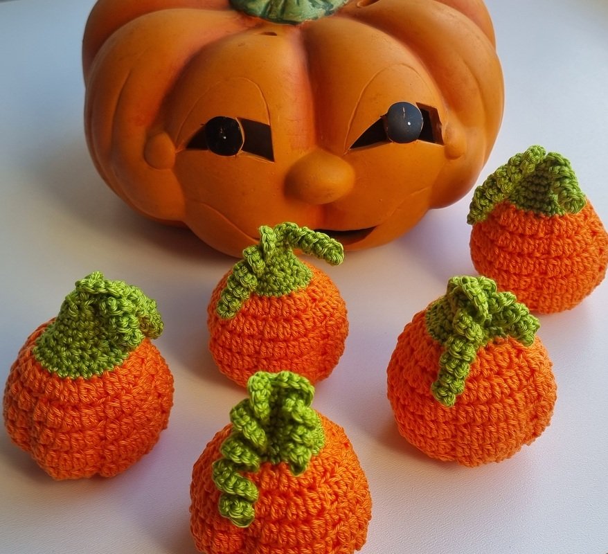 Fünf kleine gehäkelte orange Kürbisse mit grünen Blättern vor einem orangefarbenen Keramikkürbis mit Gesicht