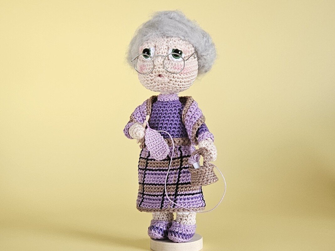 Gehäkelte Amigurumi-Oma mit grauem Haar, Brille, lila Kleidung, Häkelnadel und Garnkorb