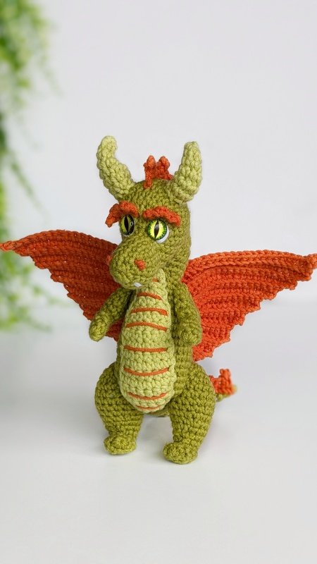 Olive-green crocheted dragon amigurumi with orange wings standing on a white surface