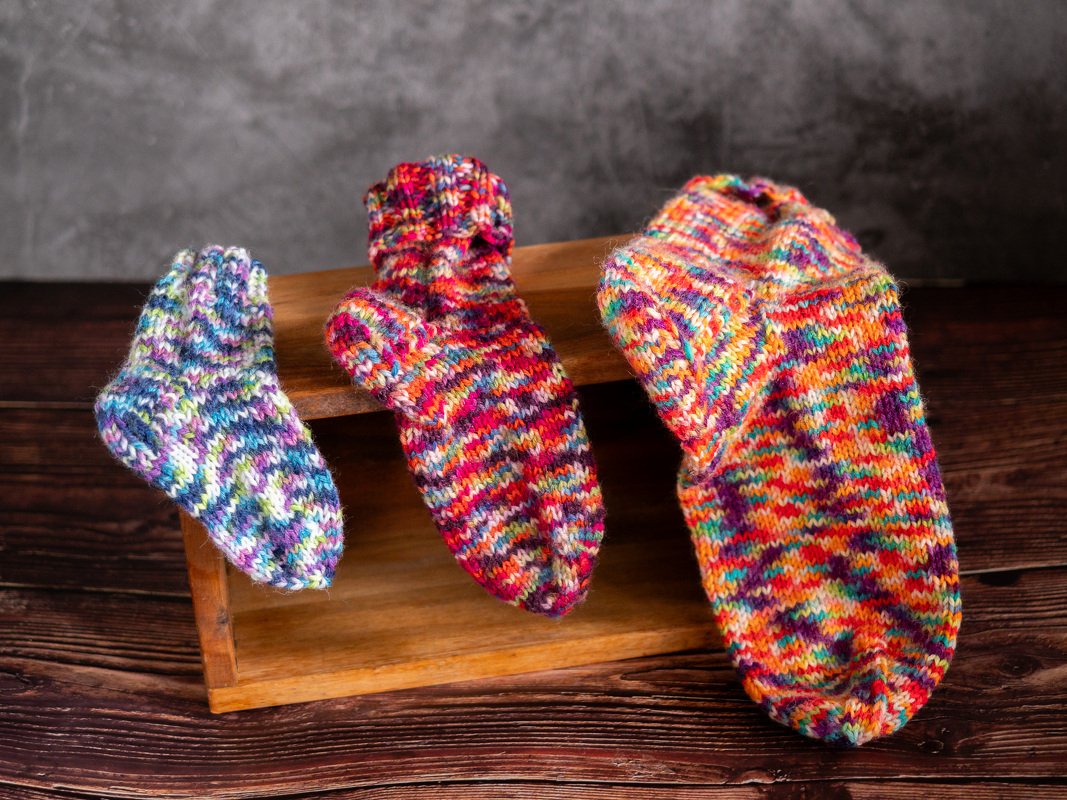 Drei handgestrickte, bunte Socken in aufsteigender Größe, drapiert auf einer Holzkiste vor neutralem Hintergrund.