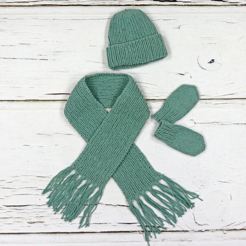 Gestricktes Baby-Set mit Mütze, Fransen-Schal und Fäustlingen auf hellen Holzplanken