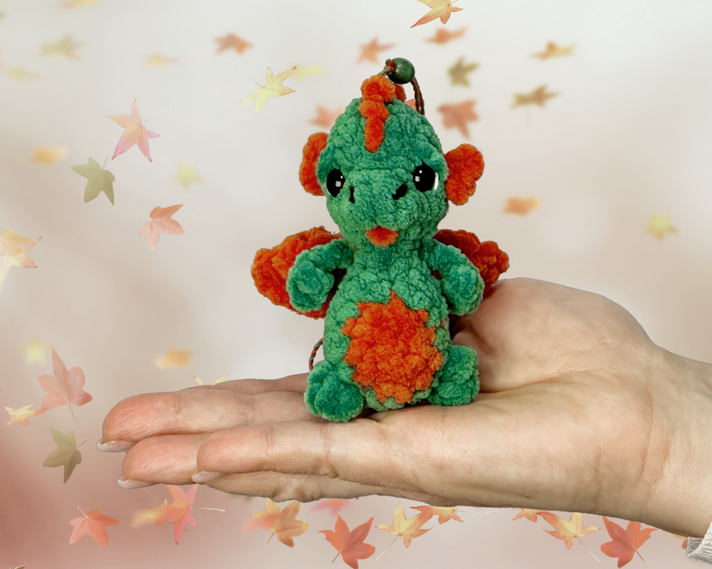 Grün-oranger gehäkelter Amigurumi-Drache vor abstraktem Herbsthintergrund, auf offener Hand platziert