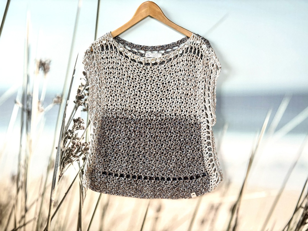 Gehäkeltes Damen‑Shirt in Creme und Braun mit Netzmuster an einem Holzbügel vor Strandkulisse.