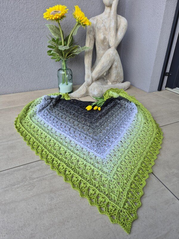 Triangle crocheted shawl in ombre from dark grey to lime green with lacy stitch and scalloped border, displayed on a tiled floor with flowers and a statue.