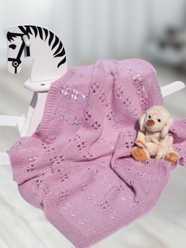 Rosa gestrickte Babydecke mit Lochmuster auf weißem Schaukelpferd neben Plüschtier