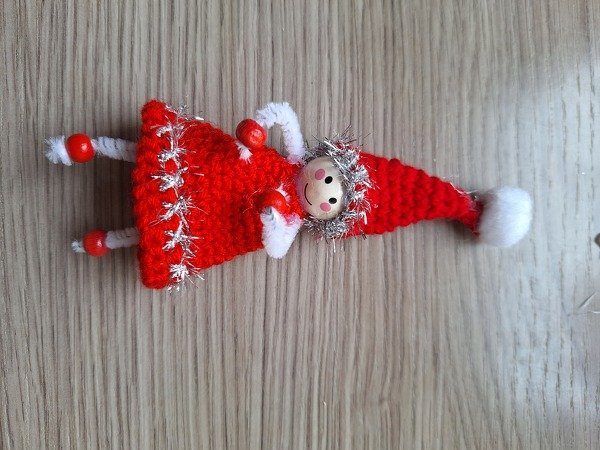 Gehäkelter roter Wichtel mit Holzkopf, silbernem Lametta und Pfeifenreiniger-Beinen auf Holzuntergrund