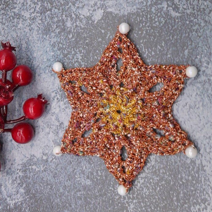 Gehäkelter sechszackiger Stern aus rotbraunem Garn mit weißen Perlen an den Spitzen, daneben rote Dekobeeren auf grauem Untergrund.