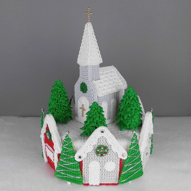 Crocheted miniature church with steepled roof and pine trees on a snowy round base
