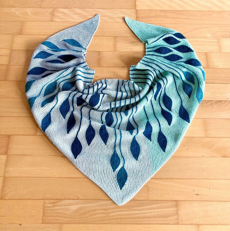 Gestricktes Dreieckstuch in kraus rechts mit blau-grünen Blattmotiven, auf hellem Holzfußboden ausgelegt