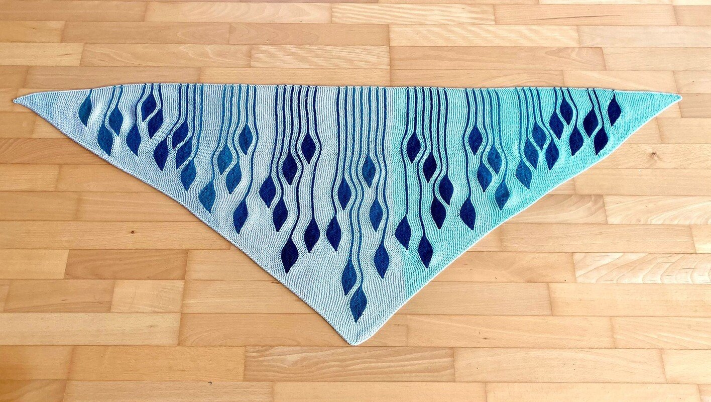 Gestricktes Dreieckstuch in Blau- und Türkistönen mit vertikalen blatt- und tropfenförmigen Mustern, auf hellem Holzfußboden liegend.