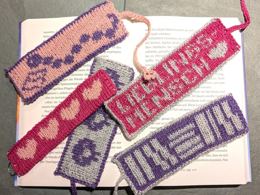 Mehrere gestrickte Lesezeichen in Rosa, Pink, Violett und Grau mit Herz- und Blockmustern auf einem aufgeschlagenen Buch