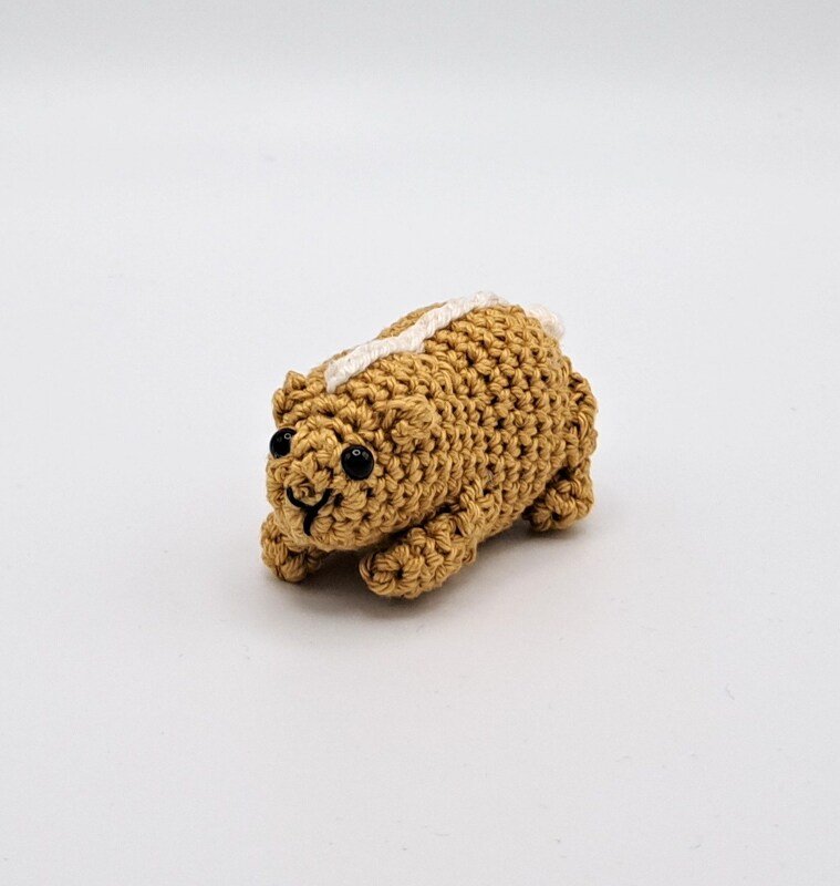 Small brown crocheted amigurumi animal with a white stripe, black safety eyes, and a stitched smile on a white background.