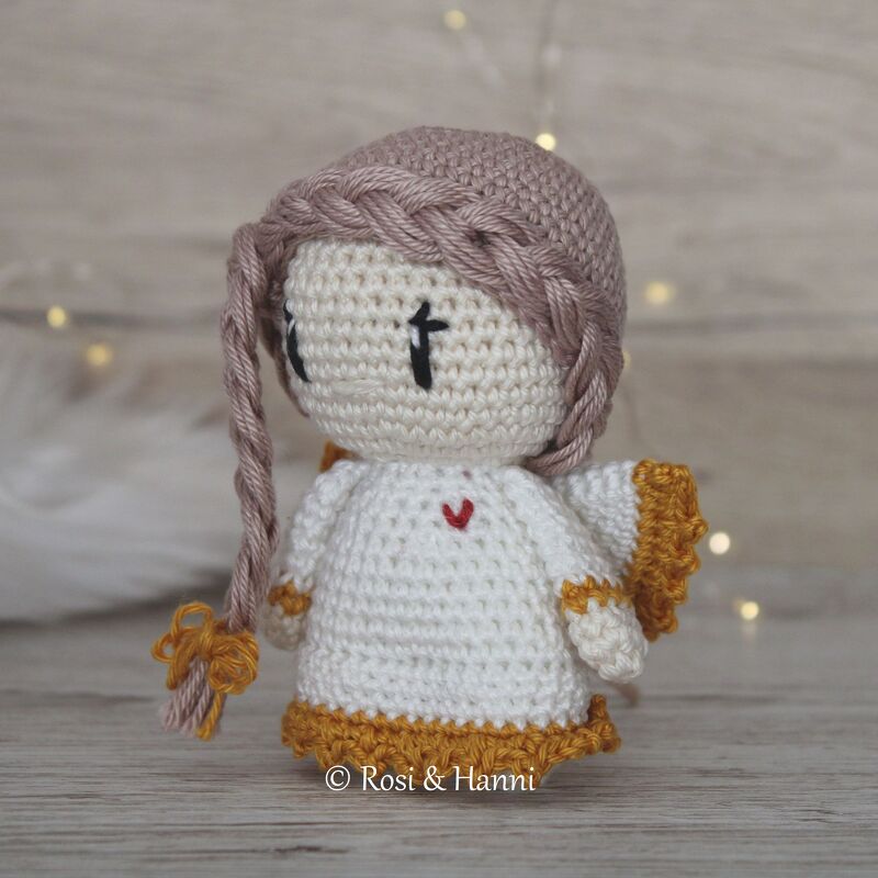 Gehäkeltes Amigurumi-Engelchen in Weiß mit hellrosa Zöpfen, senfgelben Flügeln und rotem Herz auf Holzuntergrund