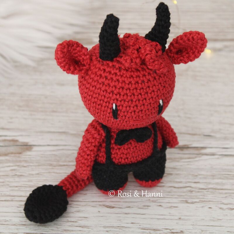 Rotes gehäkeltes Amigurumi-Teufelchen mit schwarzen Hörnern, Latzhose und Schwanz auf hellem Holz