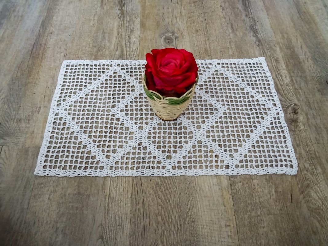 Weißes rechteckiges gehäkeltes Deckchen mit Gitter- und Rautenmuster auf Holz, rote Rose in kleinem Korb