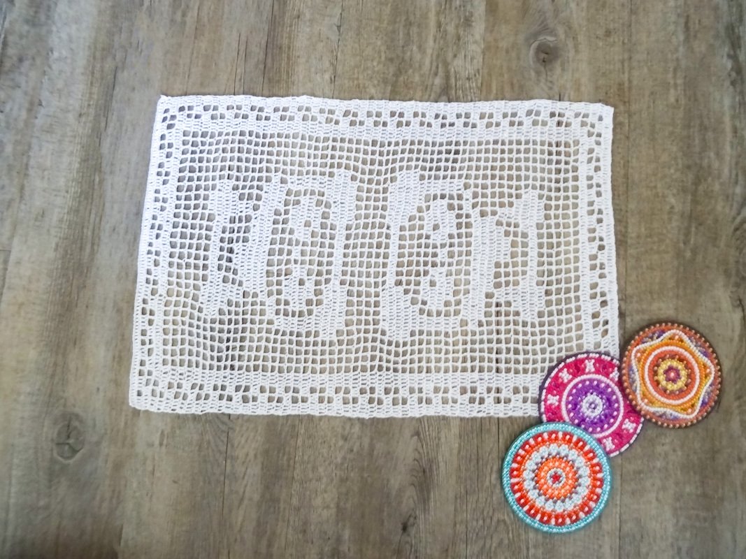 Rechteckiges weißes filetgehäkeltes Deckchen mit drei Blumenmotiven, daneben drei bunte runde Häkeluntersetzer auf Holzoberfläche
