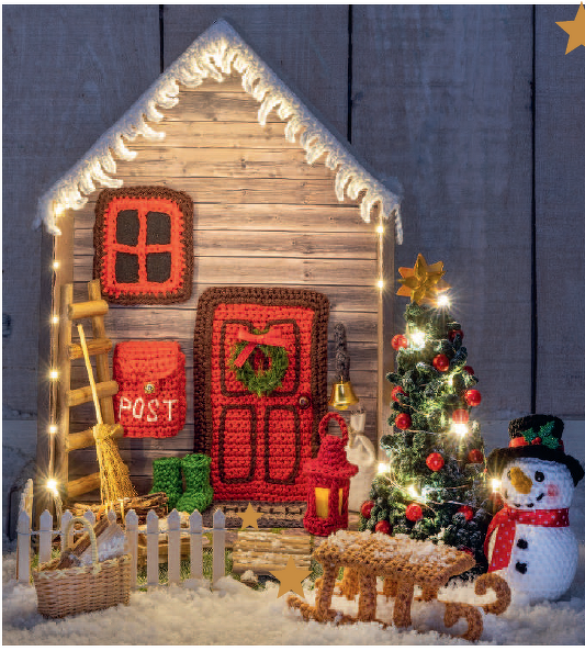 Gehäkelte Hausfassade mit roter Tür, Briefkasten, beleuchtetem Tannenbaum, Schneemann und Schlitten