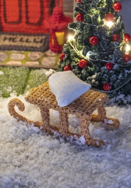 Gehäkelter Mini-Schlitten mit weißem Kissen vor beleuchtetem Weihnachtsbaum auf Kunstschnee