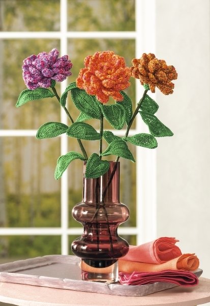 Drei gehäkelte Blumen in Violett, Orange und Rost mit grünen Blättern in dunkler Glasvase vor einem Fenster
