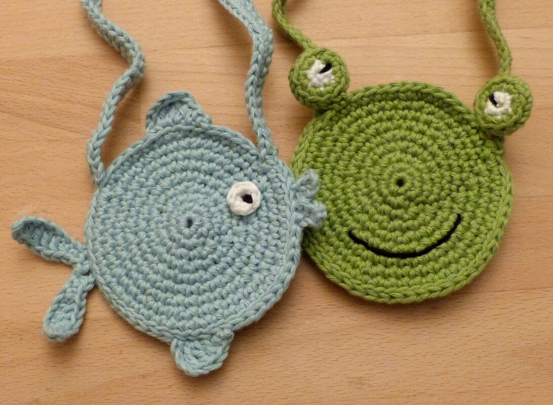 Zwei gehäkelte Kinderhandtaschen: hellblaue Fischtasche und grüne Froschtasche mit Trageriemen
