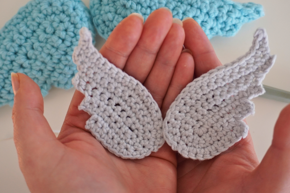 Pair of small light-gray crochet wings held in hands with blue crochet in the background.
