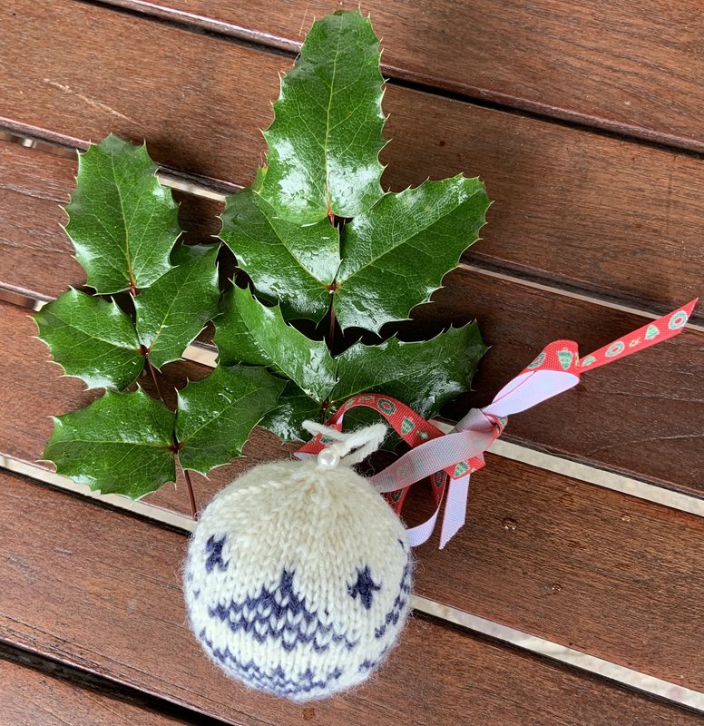 Weiße gestrickte Kugel mit blauem Muster und Schleife auf Holz neben grünen Stechpalmenblättern