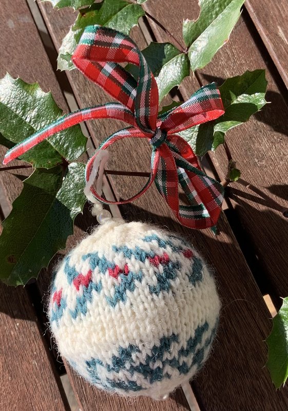 Gestrickte Weihnachtskugel in Cremeweiß, Petrol und Rot mit Fair-Isle-Muster und rot-grüner karierter Schleife.