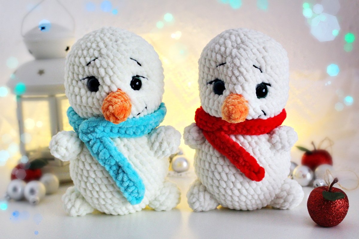Zwei flauschige, gehäkelte Schneemänner mit türkisblauem und rotem Schal vor weihnachtlichen Bokeh-Lichtern.