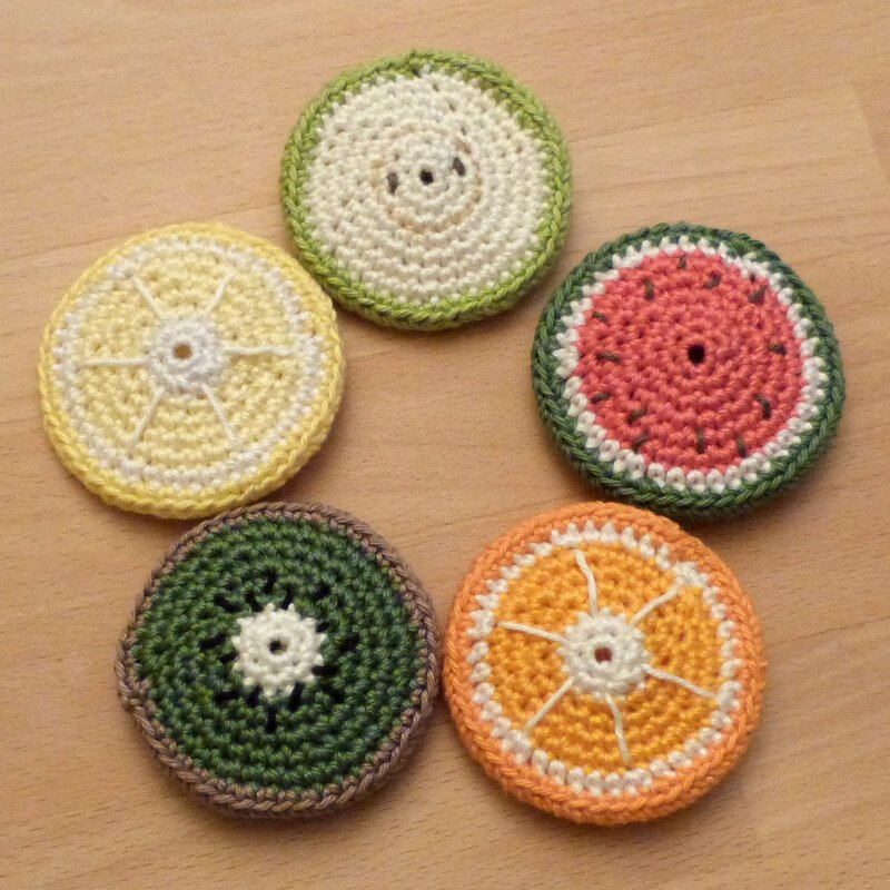Fünf gehäkelte Untersetzer in Fruchtscheiben-Optik (Zitrone, Kiwi, Orange, Wassermelone, Limette) auf hellem Holz.