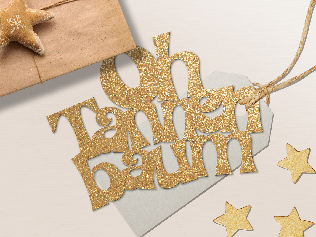 Selbstgemachter goldglitzernder 'Oh Tannenbaum'-Geschenkanhänger neben einem Päckchen und goldenen Sternen.