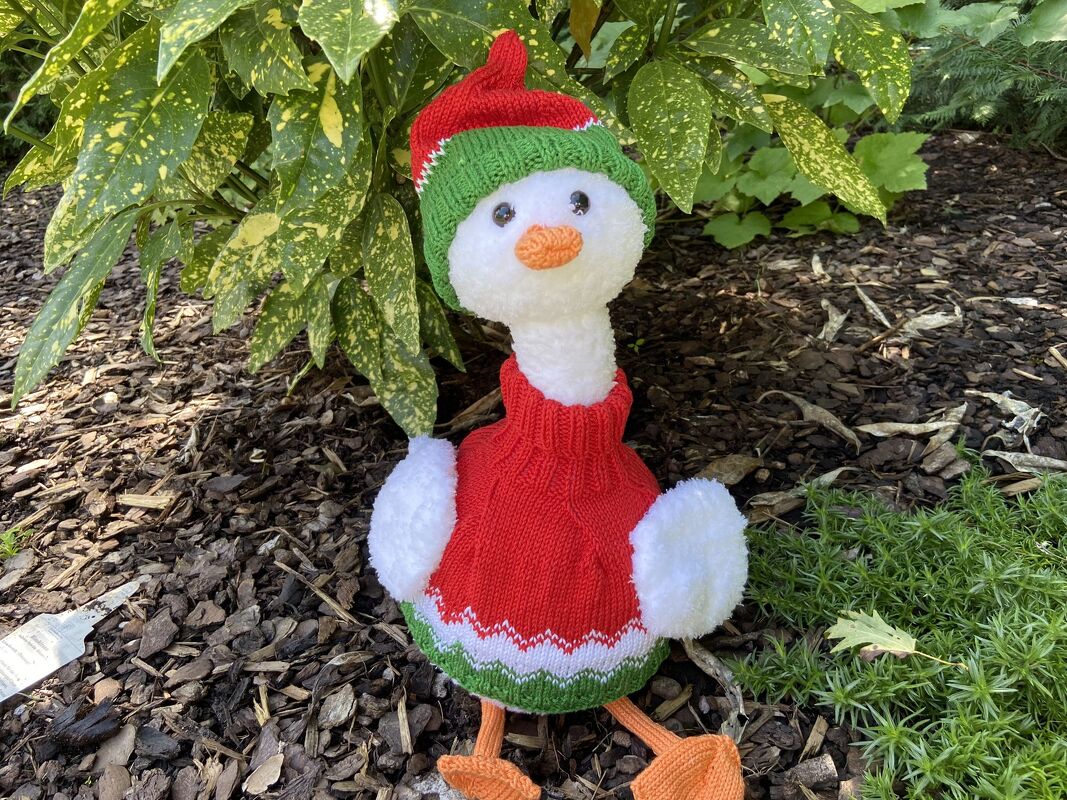 Weiß gestrickte Gans mit rotem Pullover und rot-grüner Mütze sitzend im Garten