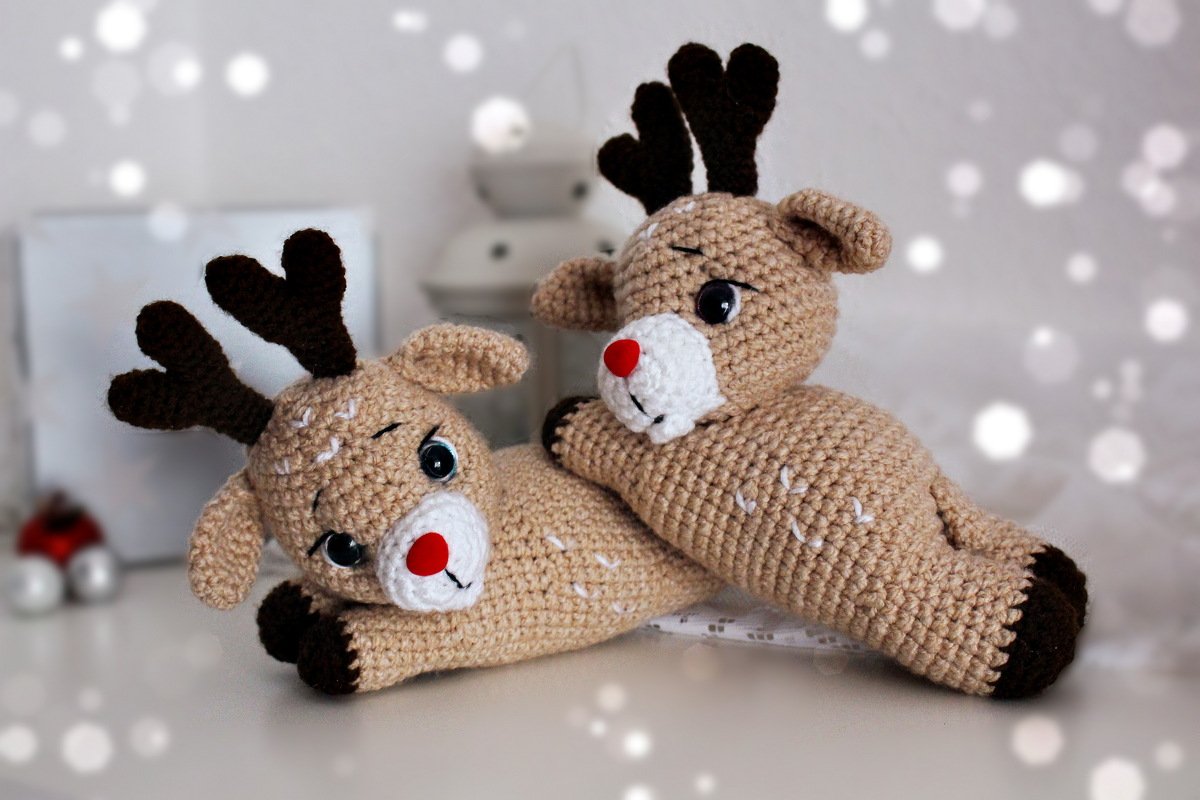 Zwei gehäkelte Amigurumi-Rentiere in Beige mit braunen Geweihen und roten Nasen, liegend