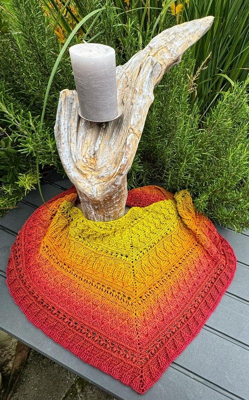 Handmade crochet triangle shawl in yellow-to-red gradient draped around a driftwood candle holder on a gray patio table