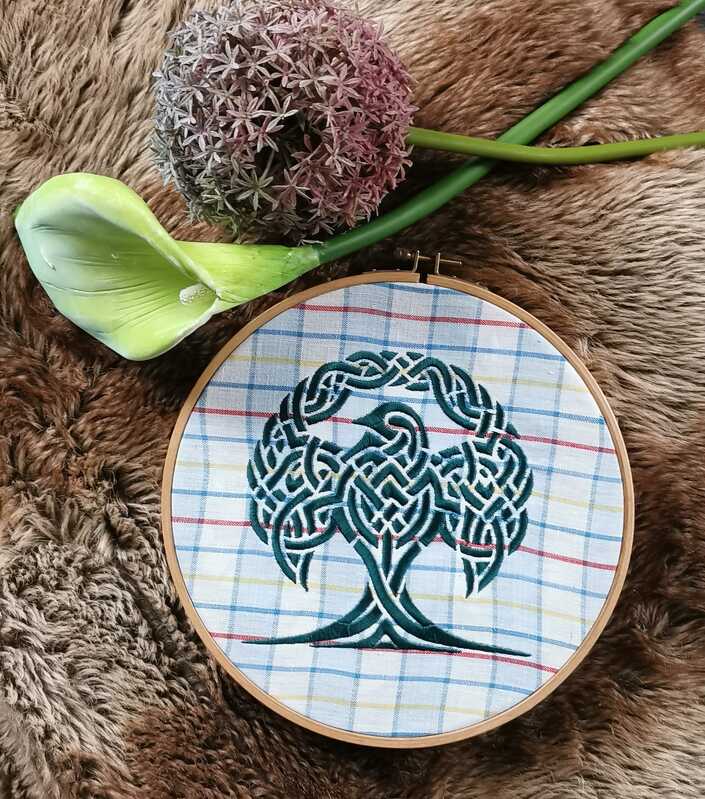 Keltischer Lebensbaum mit Vogel, gestickt auf kariertem Stoff im Holzrahmen, liegend auf braunem Fell mit Blumen.
