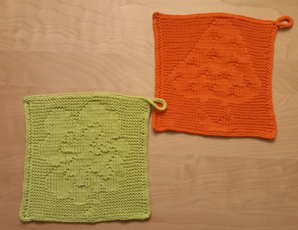 Zwei quadratische gestrickte Topflappen, hellgrün mit Kleeblattmuster und orange mit Baum-Motiv, jeweils mit Aufhängeschlaufe, auf hellem Holzuntergrund