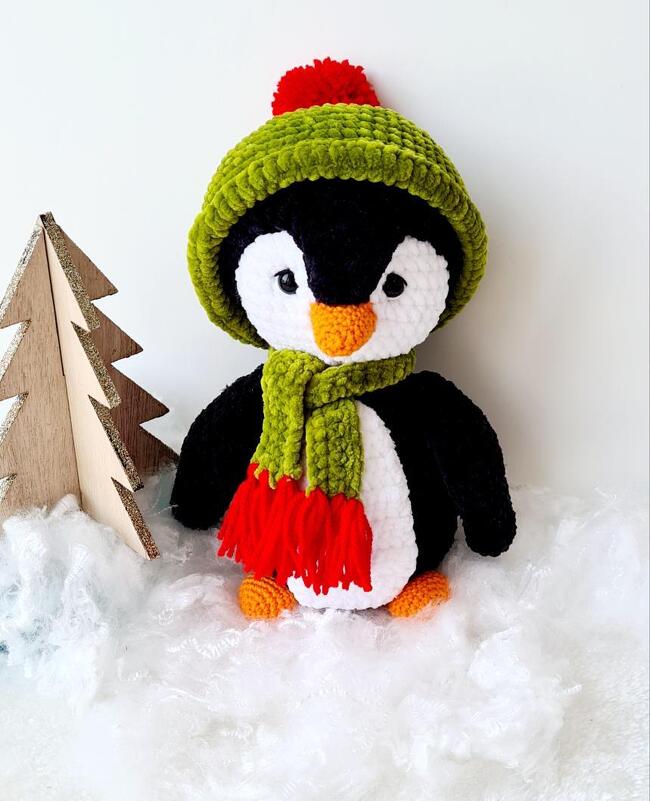 Gehäkelter Amigurumi-Pinguin mit grüner Mütze, rotem Bommel und grünem Schal, orangefarbener Schnabel und Füße