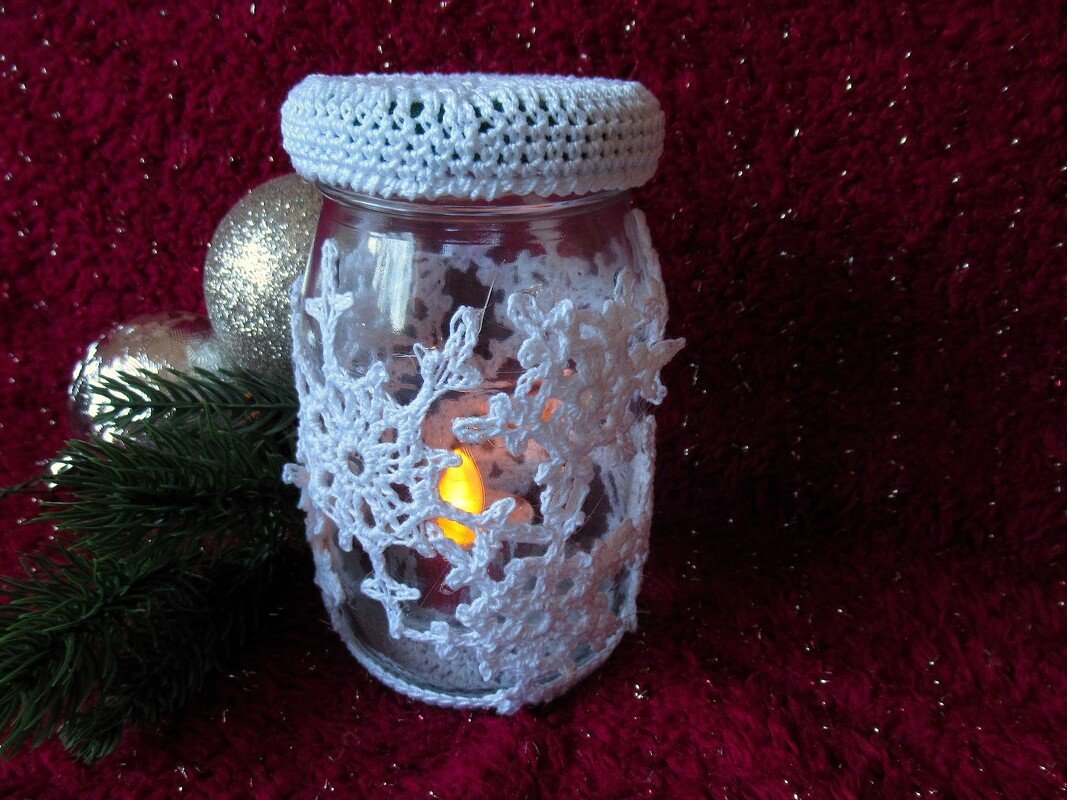 Glas-Windlicht mit weißer Häkelhülle in Schneeflockenform, Tannenzweig und silberne Weihnachtskugeln auf rotem Hintergrund