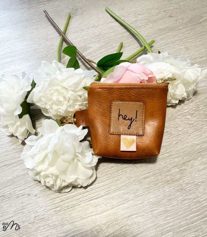 Braune Mini-Geldbörse mit 'hey!'-Patch und Herz-Applikation auf hellem Holz, umgeben von weißen Blumen