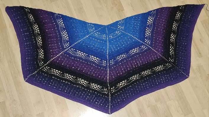 Triangular crochet shawl with blue-to-purple ombre and lace bands, laid flat on a light wooden floor.